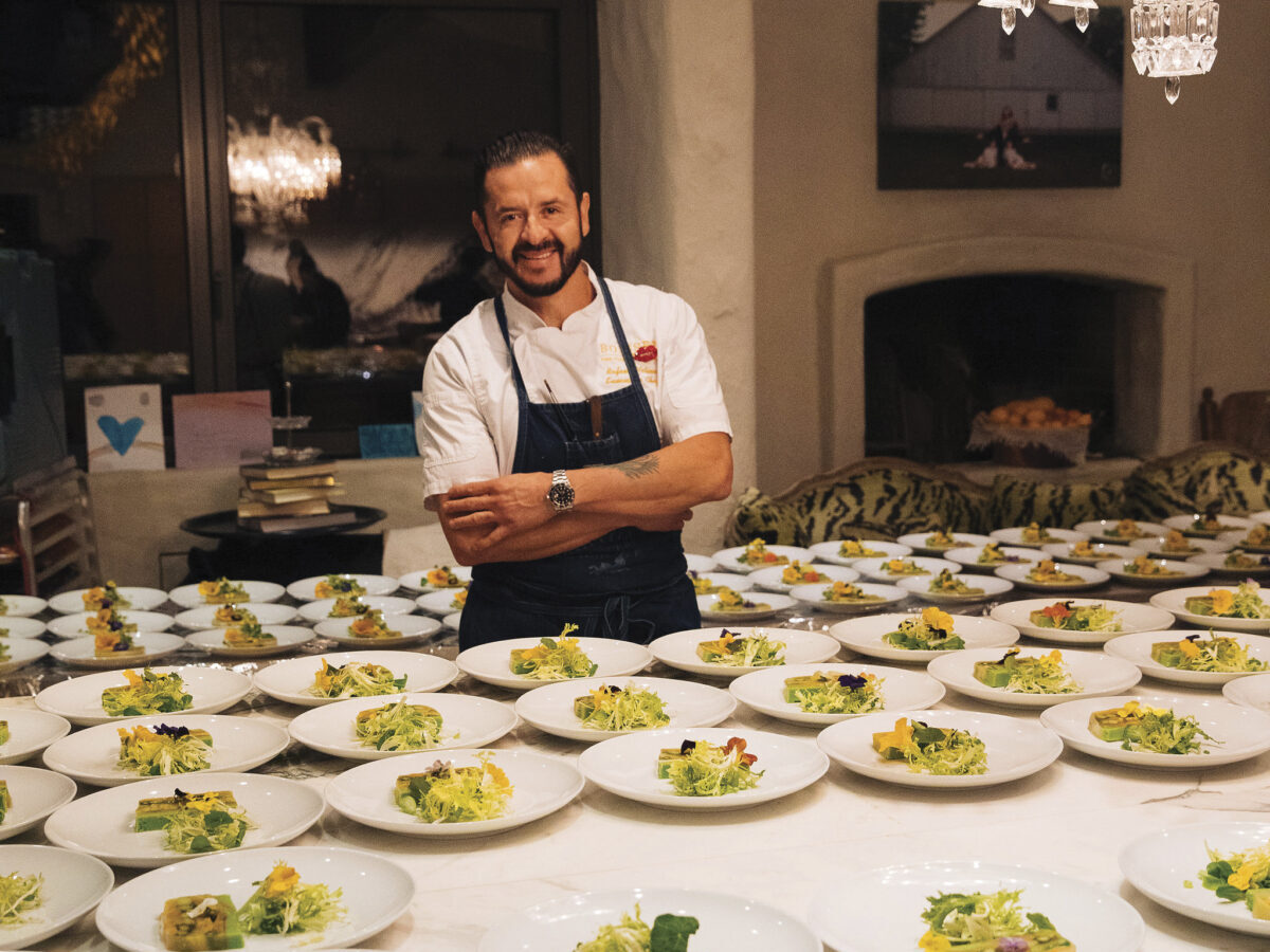 Chef Rafeal Molina standing behind a table of plated dishes in an apron and crossed arms, smiling