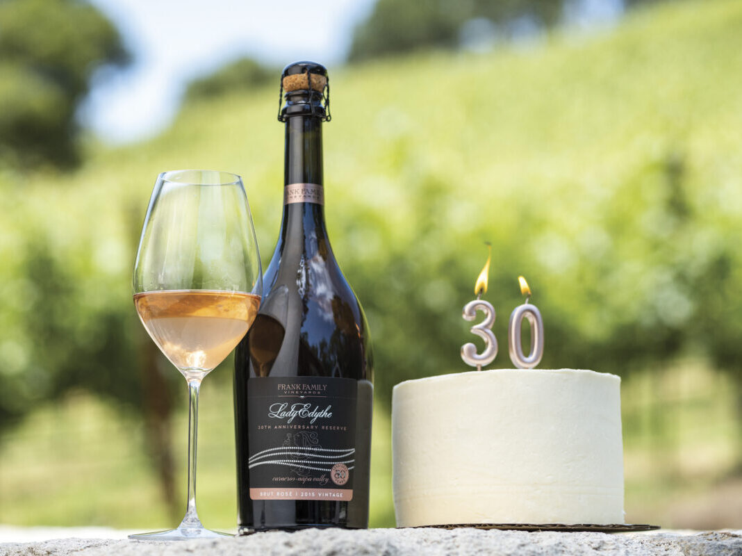 Bottle of Sparkling rose with a full glass next to a white cake with burning candles creating "30" in an outdoor setting