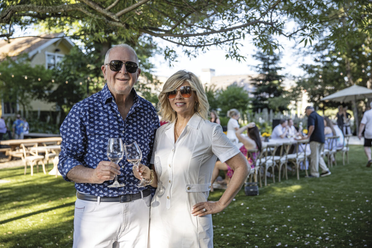 Rich and Leslie Frank, wearing sunglasses, toast each other outside in front of a tables with guests