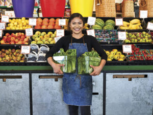 Smiling woman wearing apron and holding vegetables in front of variety of fresh fruits and vegetables