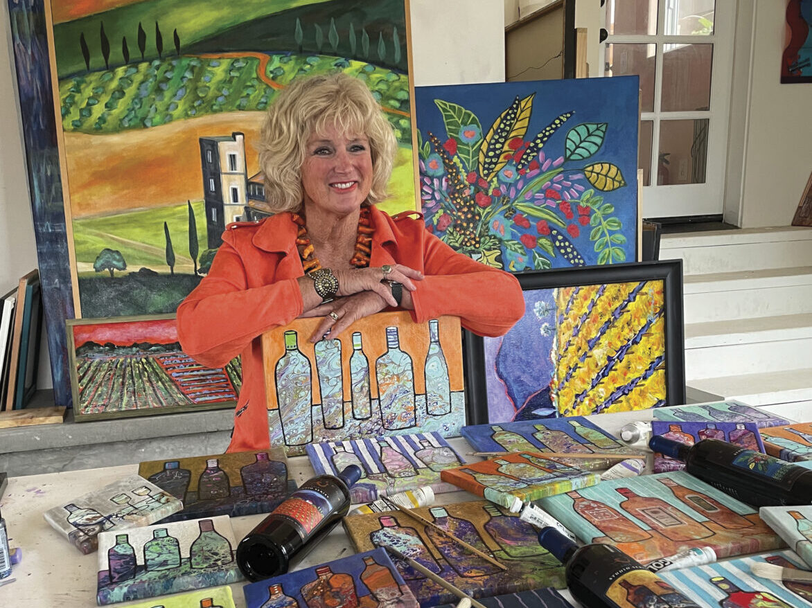 Heidi Barrett in a orange jacket posing with her colorful paintings of wine, vineyards and flowers