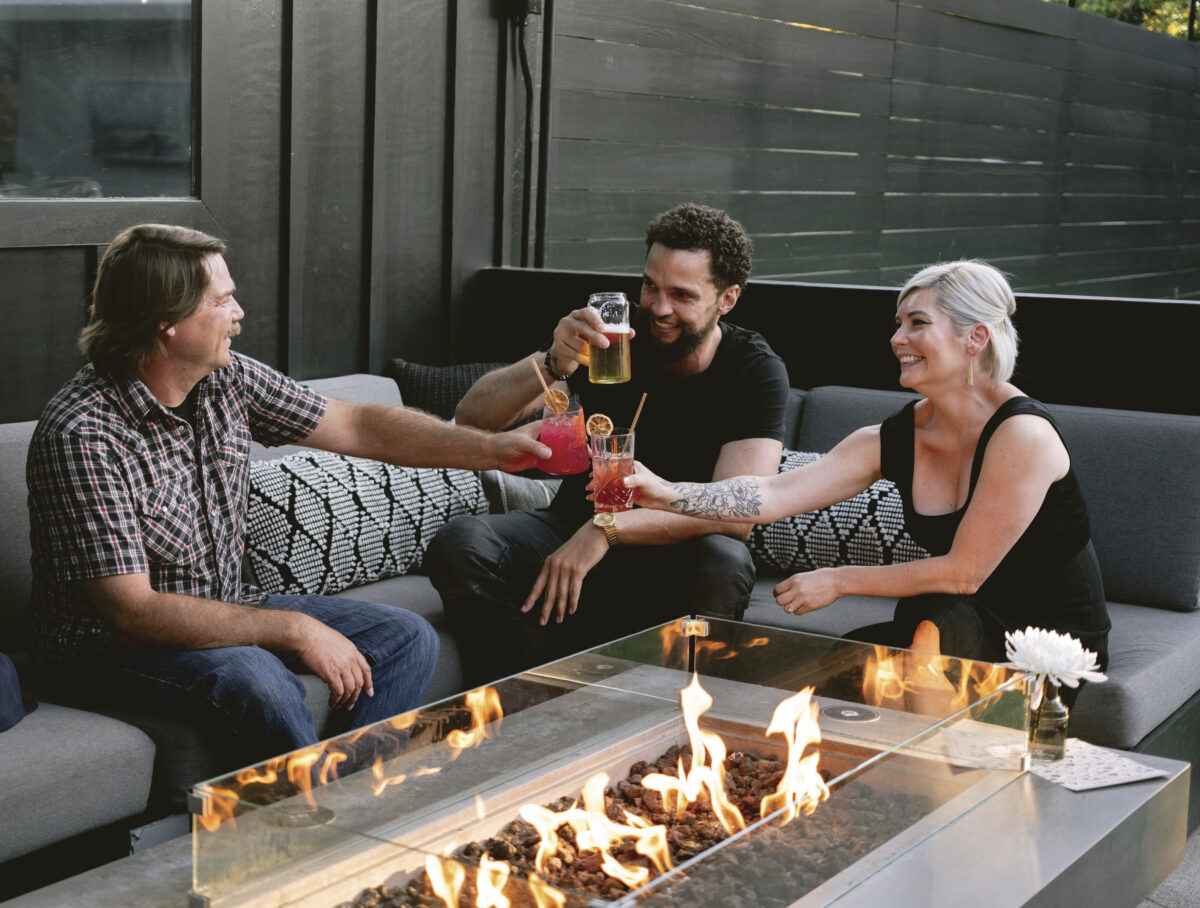 Three adults sitting outdoors at a low table with fire, toasting their adult beverages