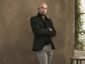 Jonathan Giacoletto in black jacket and gray plaid pants in front of brown wall with arms crossed