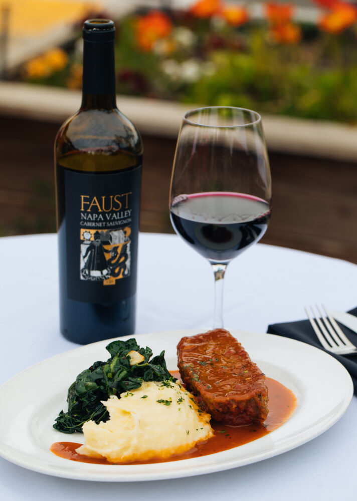 plate of meatloaf, mashed potatoes and greens with a bottle of Faust red wine and poured in wine glass on white tablecloth