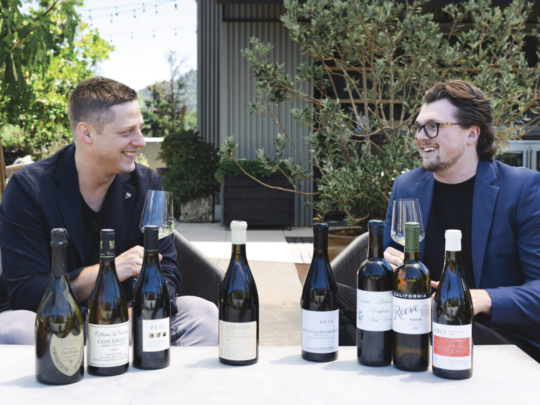 Two men sit on an outdoor couch with wine glasses and a table with various bottles of wine, smiling