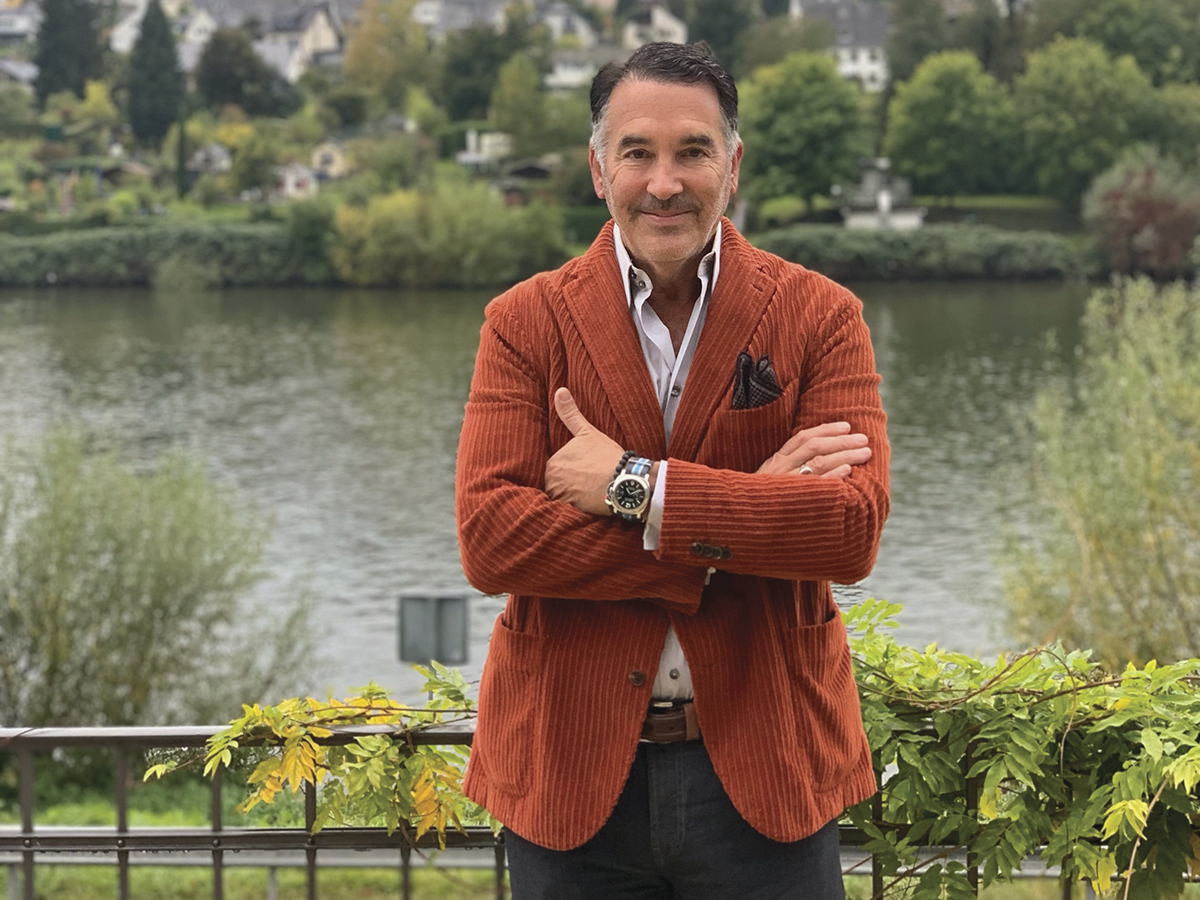 ÆRENA Gallery & Gardens founder Michael Polenske in an orange jacket with his arms crossed in front of river with greenery