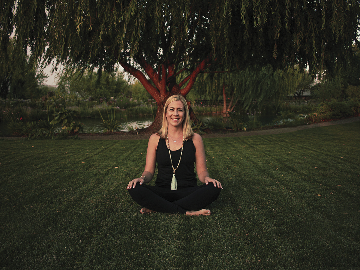 Laura Cropper of Cloud Nine Yoga, sitting outside on grass, in front of tree with crossed legs in yoga position