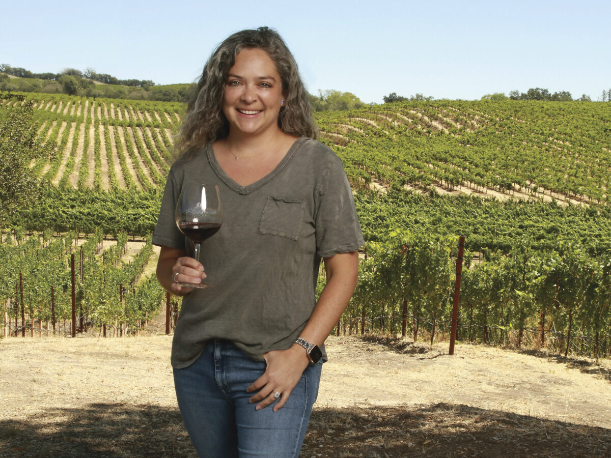 Proprietor of Enriquez Estate Winery, Cecilia Enriquez in front of their vineyard in a brown shirt and jeans, holding a glass of wine