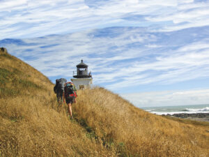 image of lighthouse on grassy hill with ocean in background with people hiking with backpacks