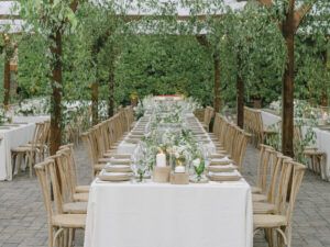 Image of long table and chairs with white tablecloth, place settings with floral arrangements under greenery on outdoor patio