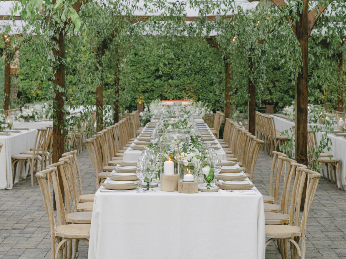 Image of long table and chairs with white tablecloth, place settings with floral arrangements under greenery on outdoor patio