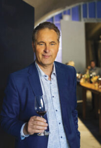 Ivo Jeramaz holding a glass of red wine in a blue suit jacket and light blue shirt