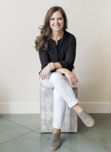 portrait of Kristin Maher of Napa Farmhouse Interiors with wearing white pants, black shirt, seated with concrete floors and neutral backdrop