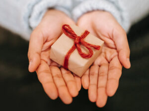 two hands with palms facing up holding a small gift wrapped box with red bow
