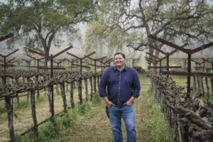 Garrett Buckland standing in vineyard with hands in pockets wearing a navy shirt and jeans