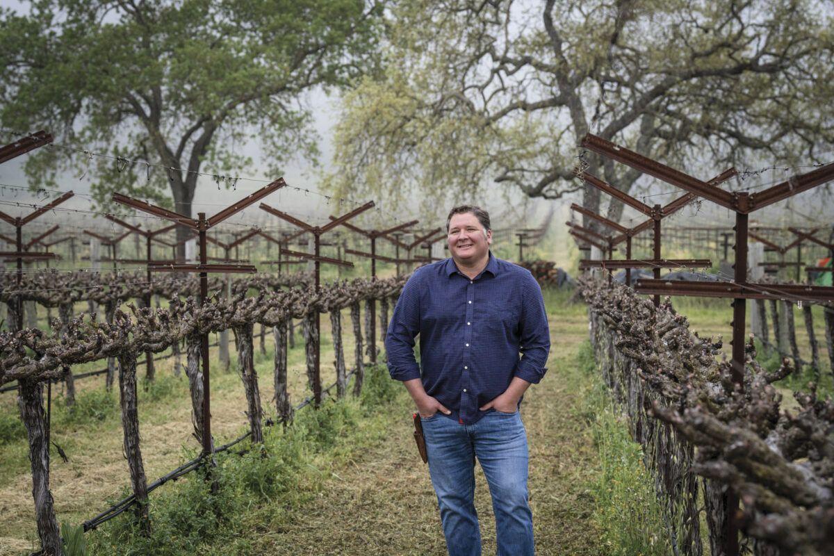 Garrett Buckland standing in vineyard with hands in pockets wearing a navy shirt and jeans