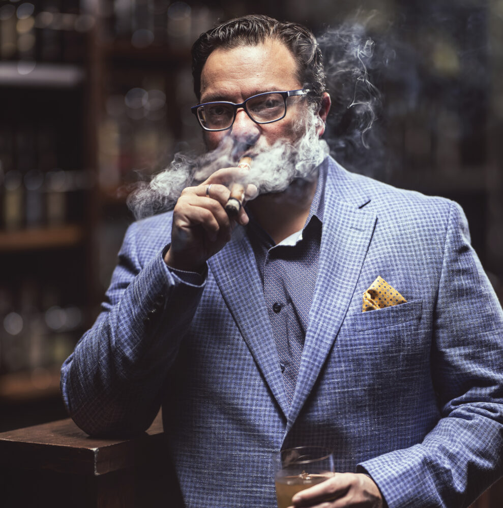 Eric Smaldino smoking cigar with drink in hand wearing a blue suit and glasses