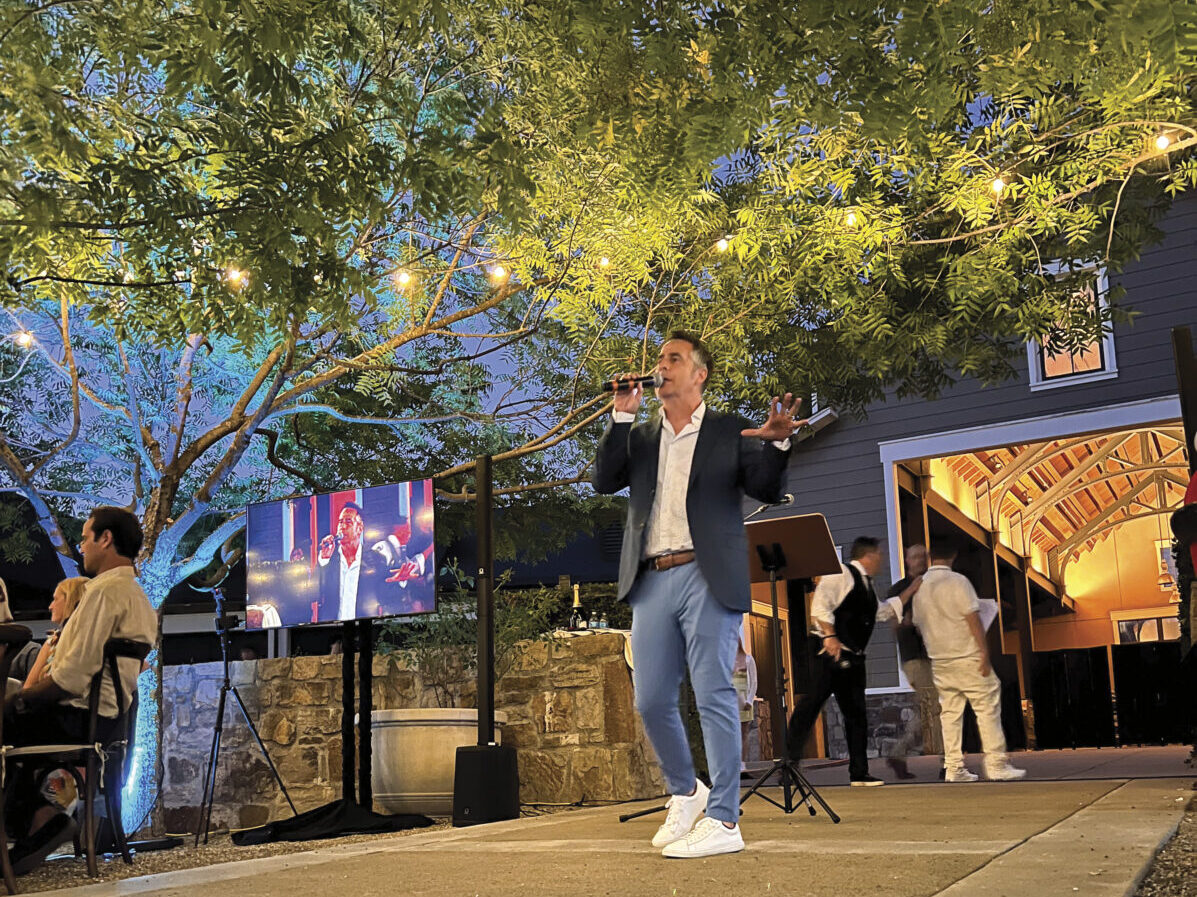 Trent Yaconelli hosting the BIG Night LIVE Auction and Gala in Napa, standing in jeans and navy blazer, speaking into a microphone outside under a tree with string lights in the evening // Photo by Danny Hernandez