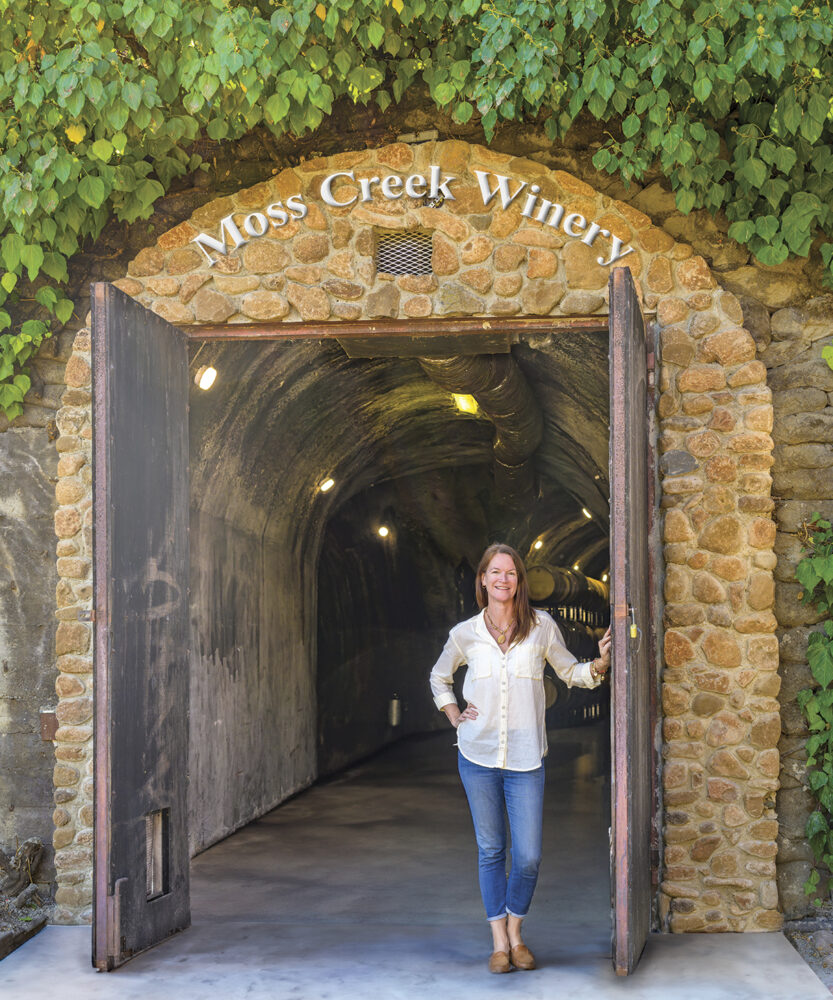 Claire Hobday standing in archway of brick and vine covered entrance of Moss Creek Winery 