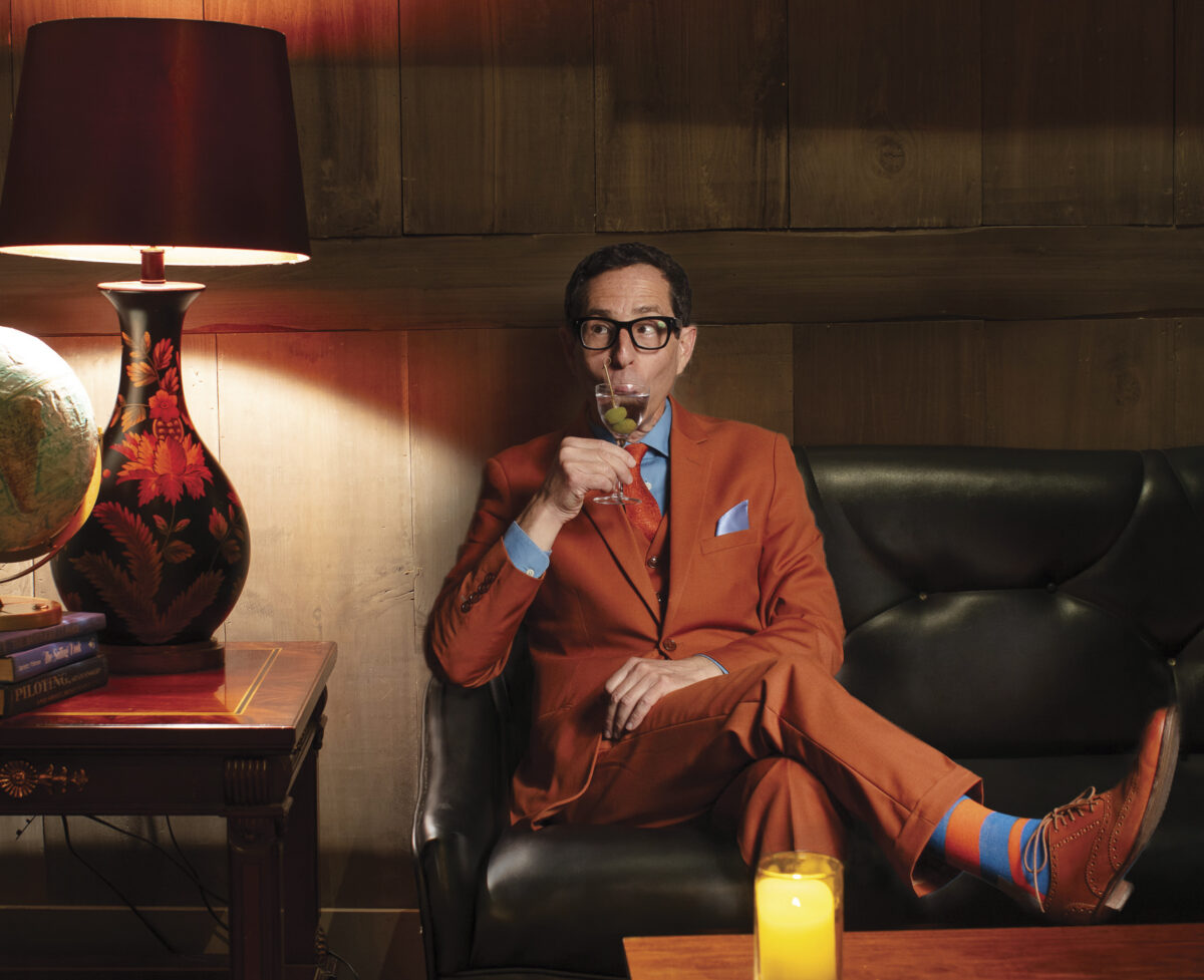 Judd Finklestein seated on a couch in a dark room wearing an orange suite sipping wine