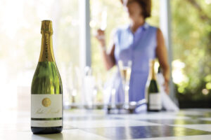 Woman in purple shirt holding a glass of sparkling wine with bottles of Paula Kornell sparkling wine in the foreground on a table