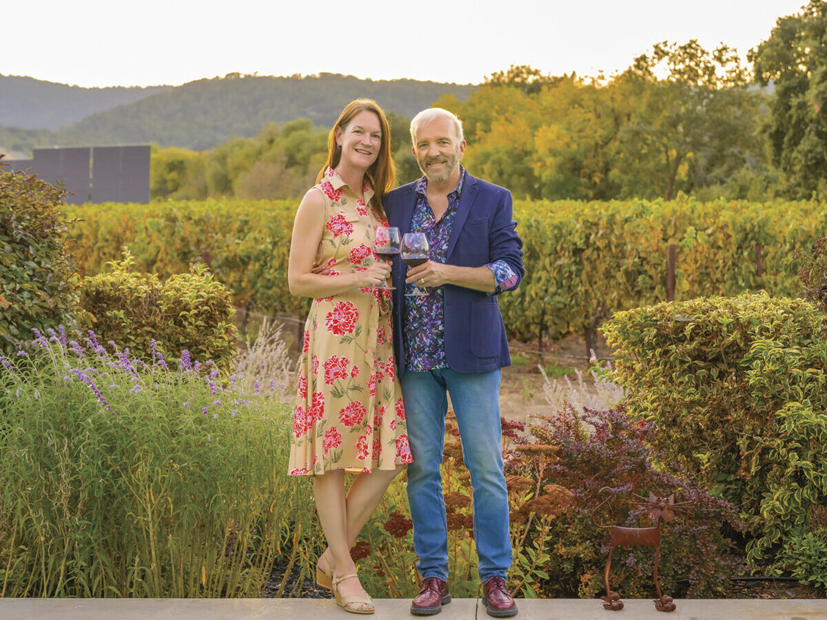 Claire Hobday and Damian Archbold stand in front of garden with mountain in distance, holding glasses of red wine