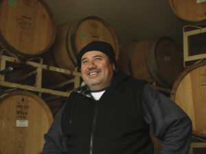Miguel Caratachea in black hat, black smiling with wood wine barrels behind him