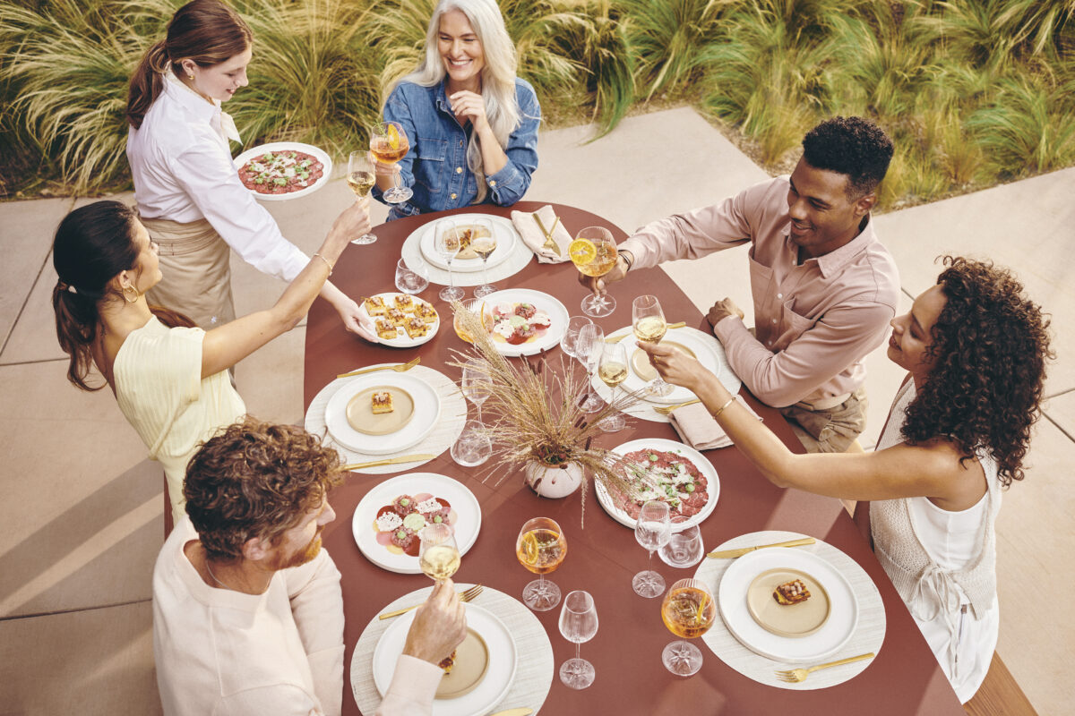 group of 4 smiling adults seated at outdoor table with plates of food, raising glasses for a toast with waitress delivering plates of food