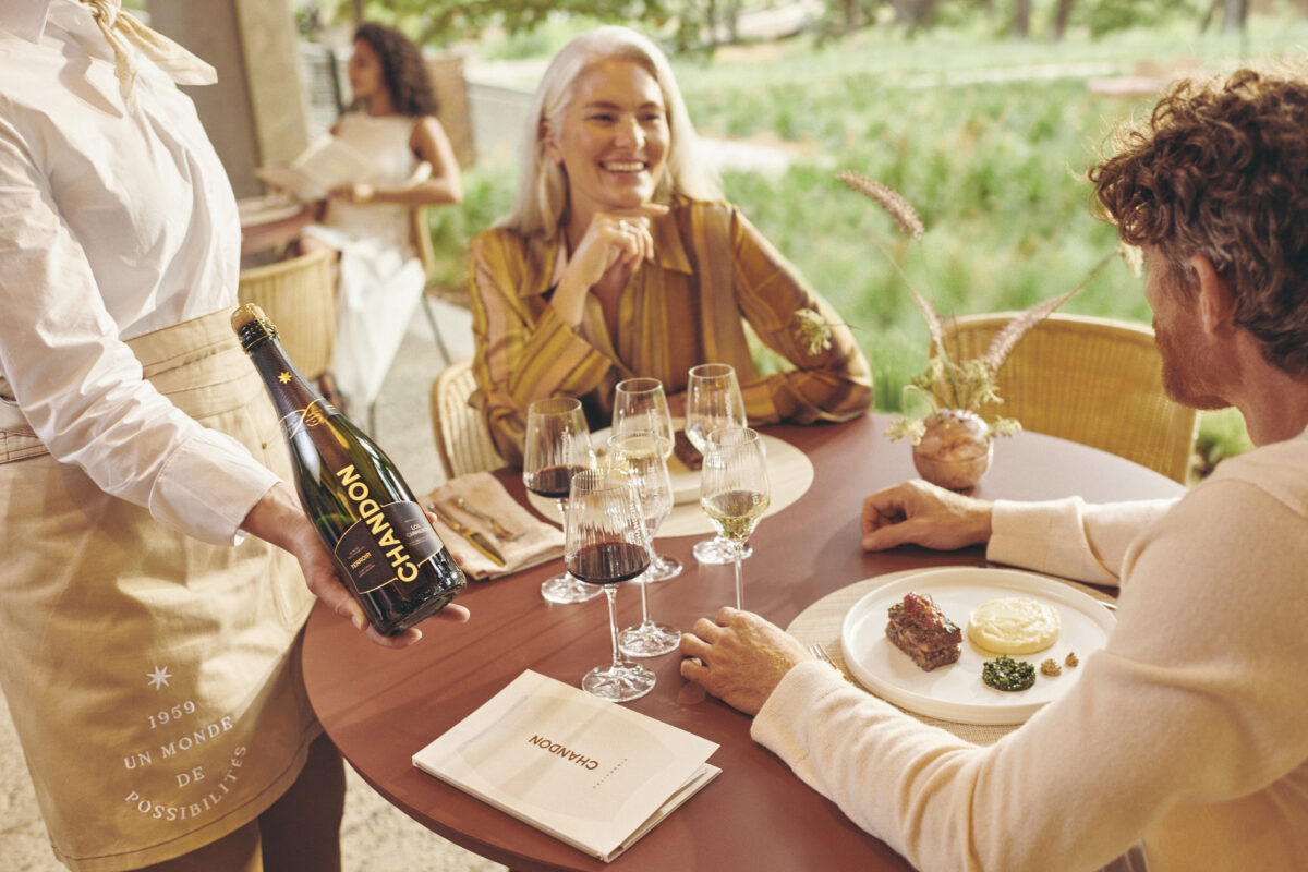 man and woman seated at outdoor table with plates of food with glasses lined up for a wine tasting with waitress offering a bottle of Chandon sparkling wine