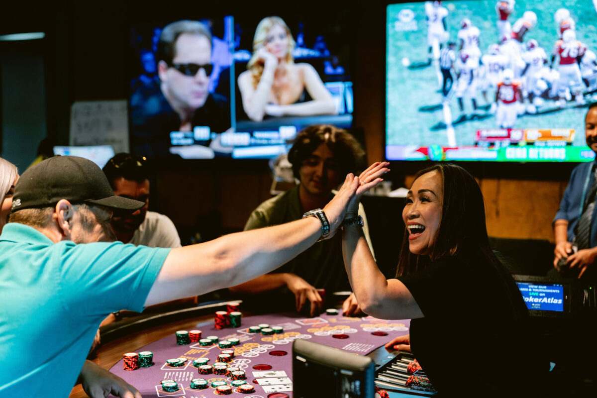 Man and woman at casino talbe, high-fiving and smiling with tv screens displaying sports and poker in background