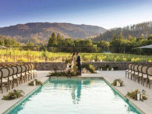Bride and groom kissing by swimming pool, setup chairs for guests at their wedding and vines and mountains in the background