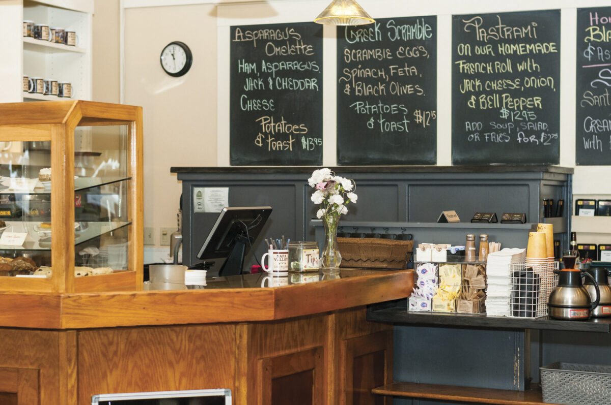 wooden counter of gillwoods cafe with a case of baked goods and computer screen with chalkboard in background with food selections written on the board. Coffee station with cups and flower in vase are also in view.