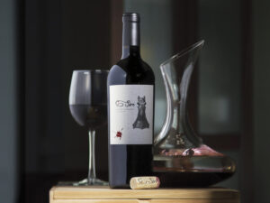 bottle of Sire wine on wooden box with poured red wine in glass, glass decantor and cork with dark background