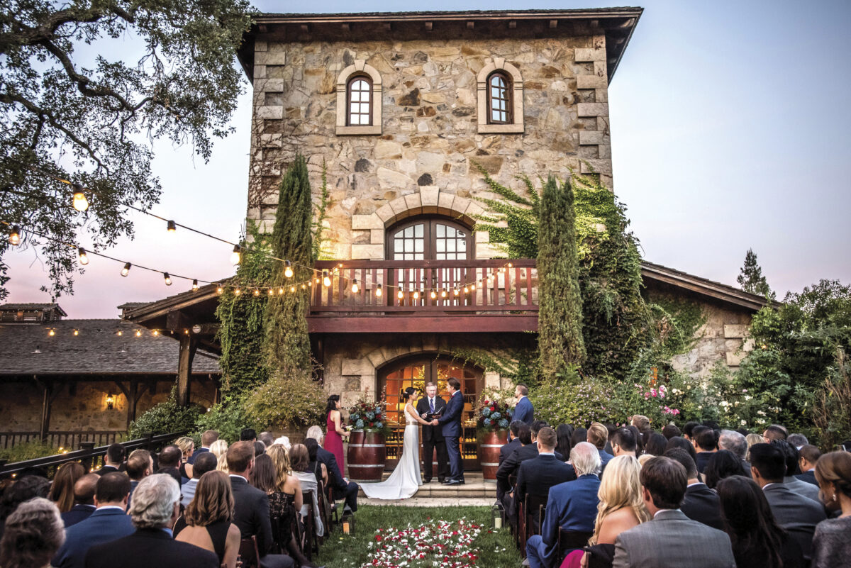 outdoor wedding during the day in front of V. Sattui winery, a stone tall building with landscaped courtyard where bride, groom and guests have the ceremony