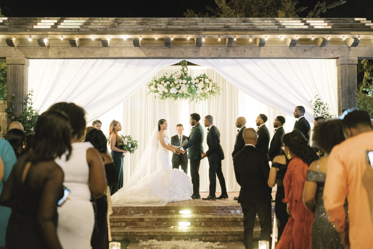 image of a wedding ceremony at Estate Yountville, set indoors with bride, groom holding hands amongst their wedding party with wedding guests looking on 