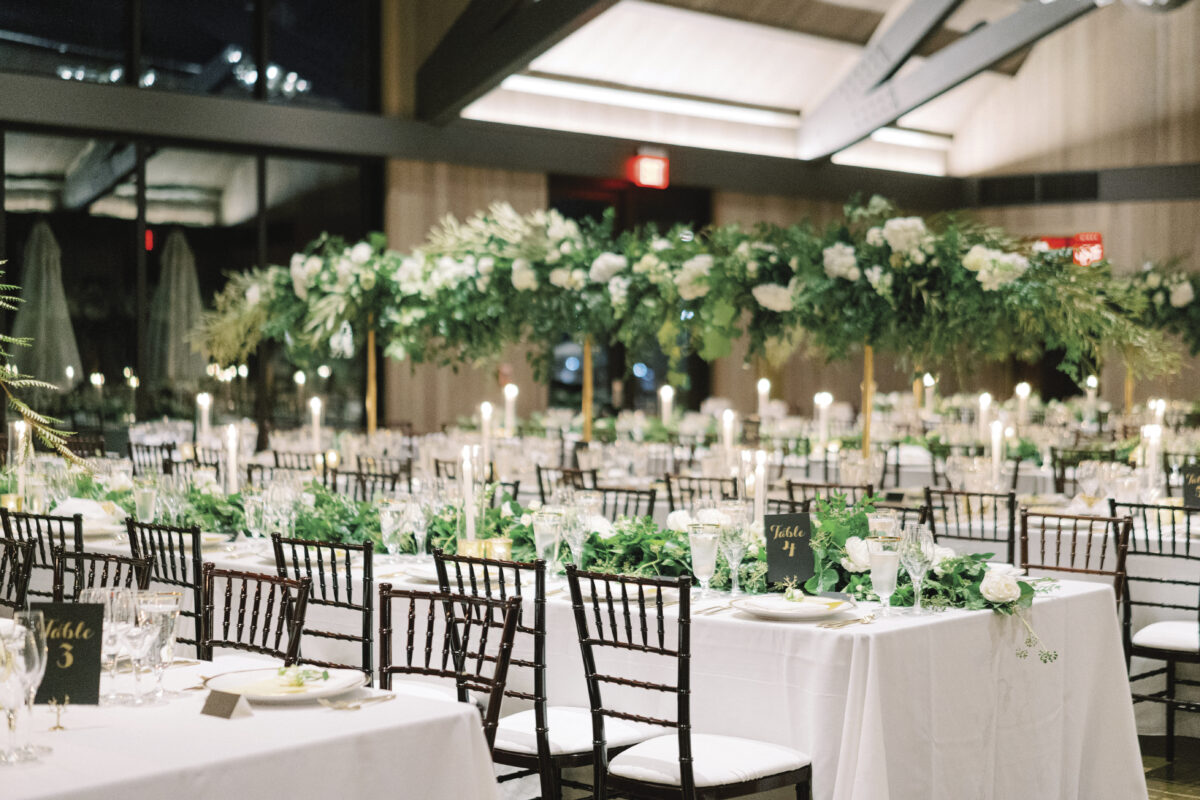 long rectangular tables set indoors with white tablecloths, greenergy, table numbers chairs and floral trees in background