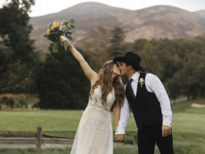 Bride holds her bouquet of flower up and kisses the groom with a cowboy hat in front of maintains at Napa Valley Country Club
