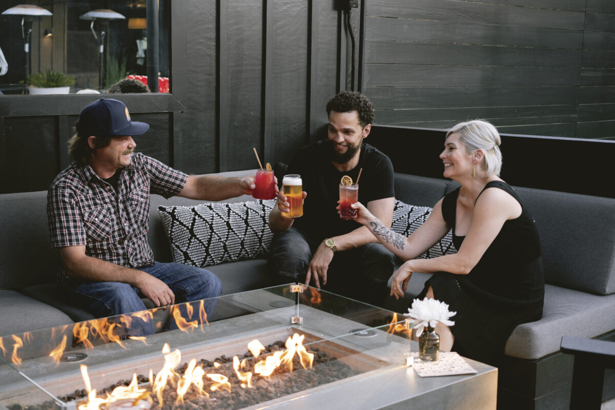 three adults sitting outside on couches on the patio of The Lincoln near a fire, toasting and smiling