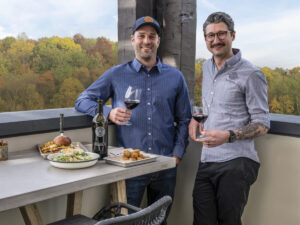 Priest Ranch Winemaker, Cody Hurd and Chef Don Solomon on the patio countertop with plates of food and a bottle of wine at the Kitchen at Priest Ranch with blue sky and trees behind them.
