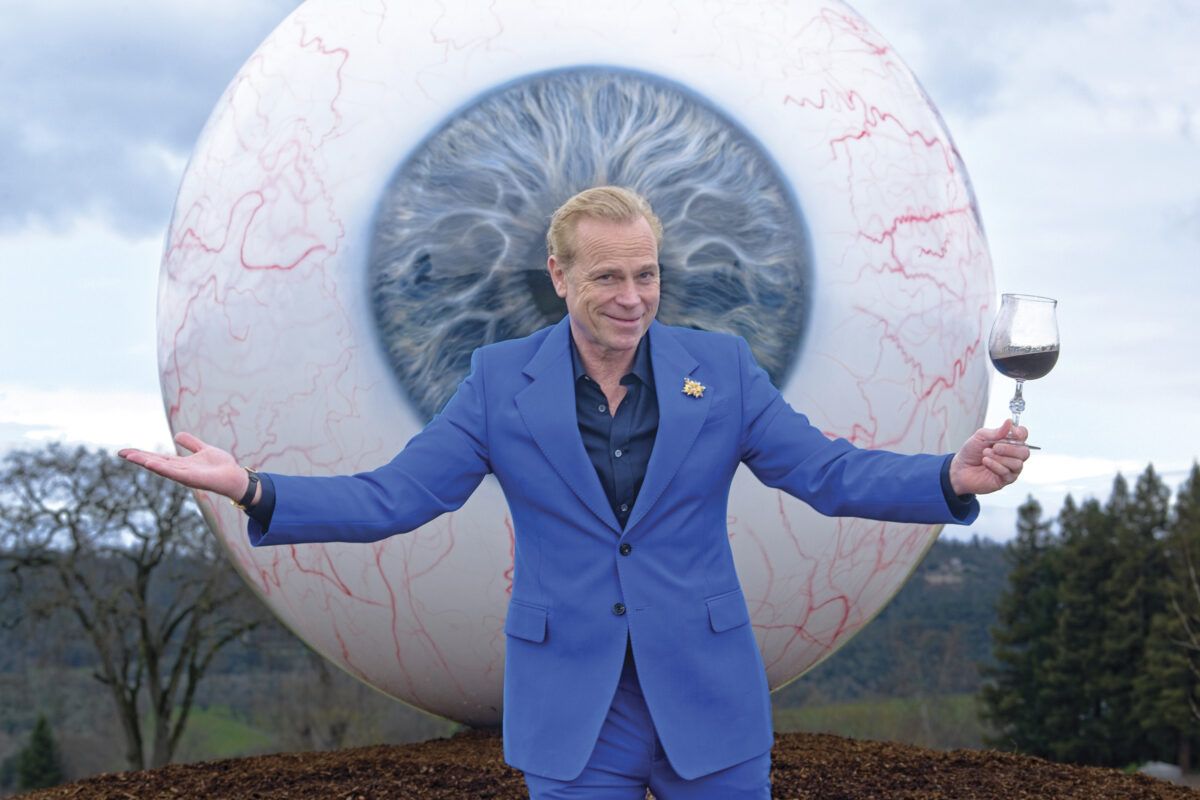 Jean-Charles Boisset standing in blue suit, holding a glass of red wine in front of a sculpture of a large eyeball