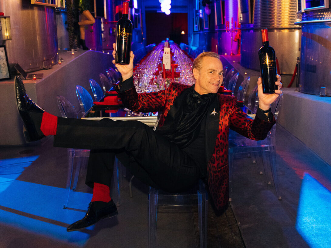 Jean-Charles Boisset sitting in chair with leg kicked up, wearing red socks and jacket with and black shoes and suit, smiling, holding 2 bottles of wine in a moody room with couches, table, chairs and chandelier, lit in blues, purples and pinks