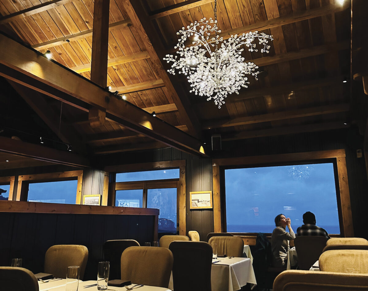 Interior of dining room with large chandelier, wooden ceiling, white tablecloth tables and chairs, with ocean view