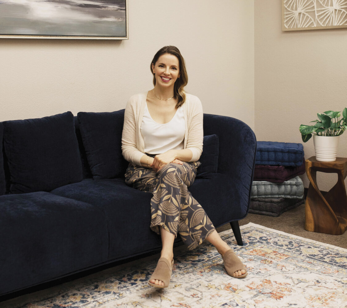 woman sitting on navy couch with leg crossed, smiling with plant and art on wall