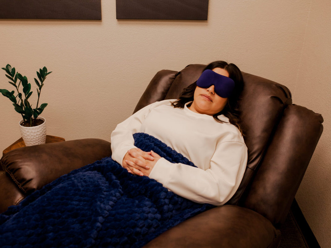 woman relaxing on leather recliner chair, with blue eye mask over her eyes, hands crossed on her lap, with blue blanket covering her in room with plant and wall art