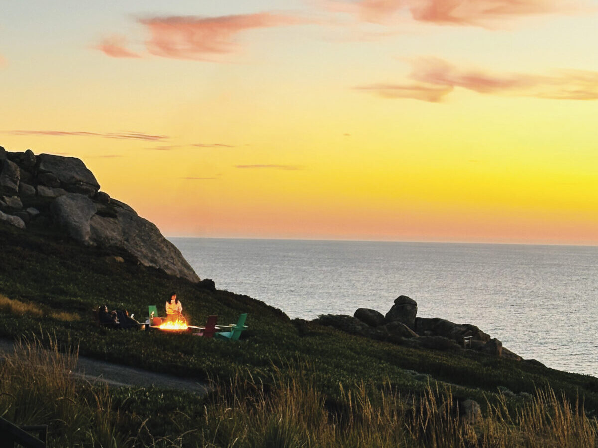 view of bonfire on grassy, rock cliff, oceanside with people sitting in chairs around it at sunset with ocean behind