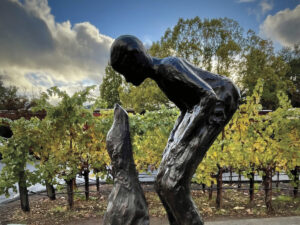 bronze sculpture of person bending looking at dog in front of vineyard and blue sky