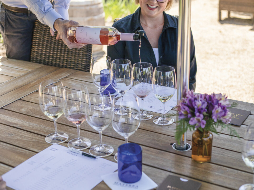 woman sitting at outdoor wooden table with wine glasses lined up for tasting, with waiter pouring rose into glass. Purple flowers and blue water glasses are also on the table