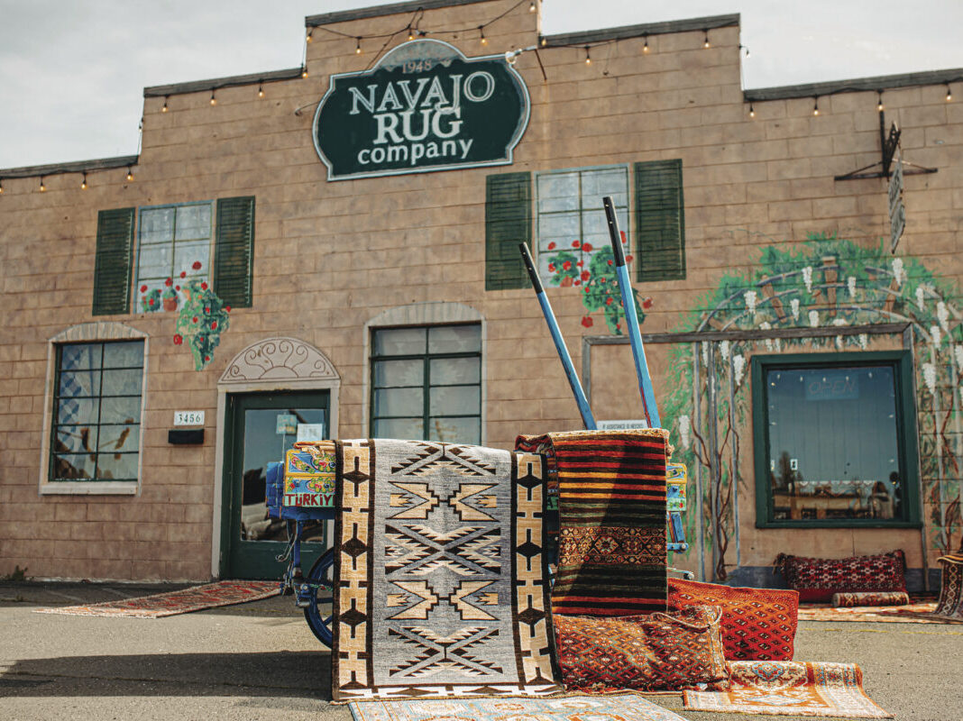 exterior of Navajo Rug Company brick building with green shutters on windows with several rugs displayed out front