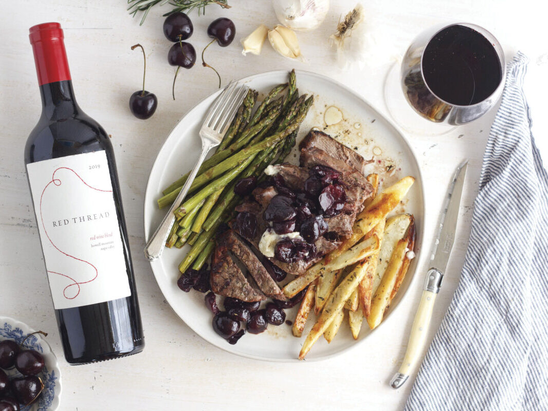 bottle of red thread sine, glass of red wine and plate of food including asparagus, french fries and meat on table with white tablecloth