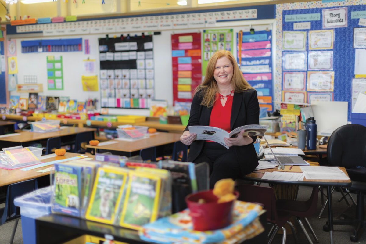 Jennifer Stewart of Napa Valley Education Foundation seated on school desk with open book in brightly colored classroom, smiling